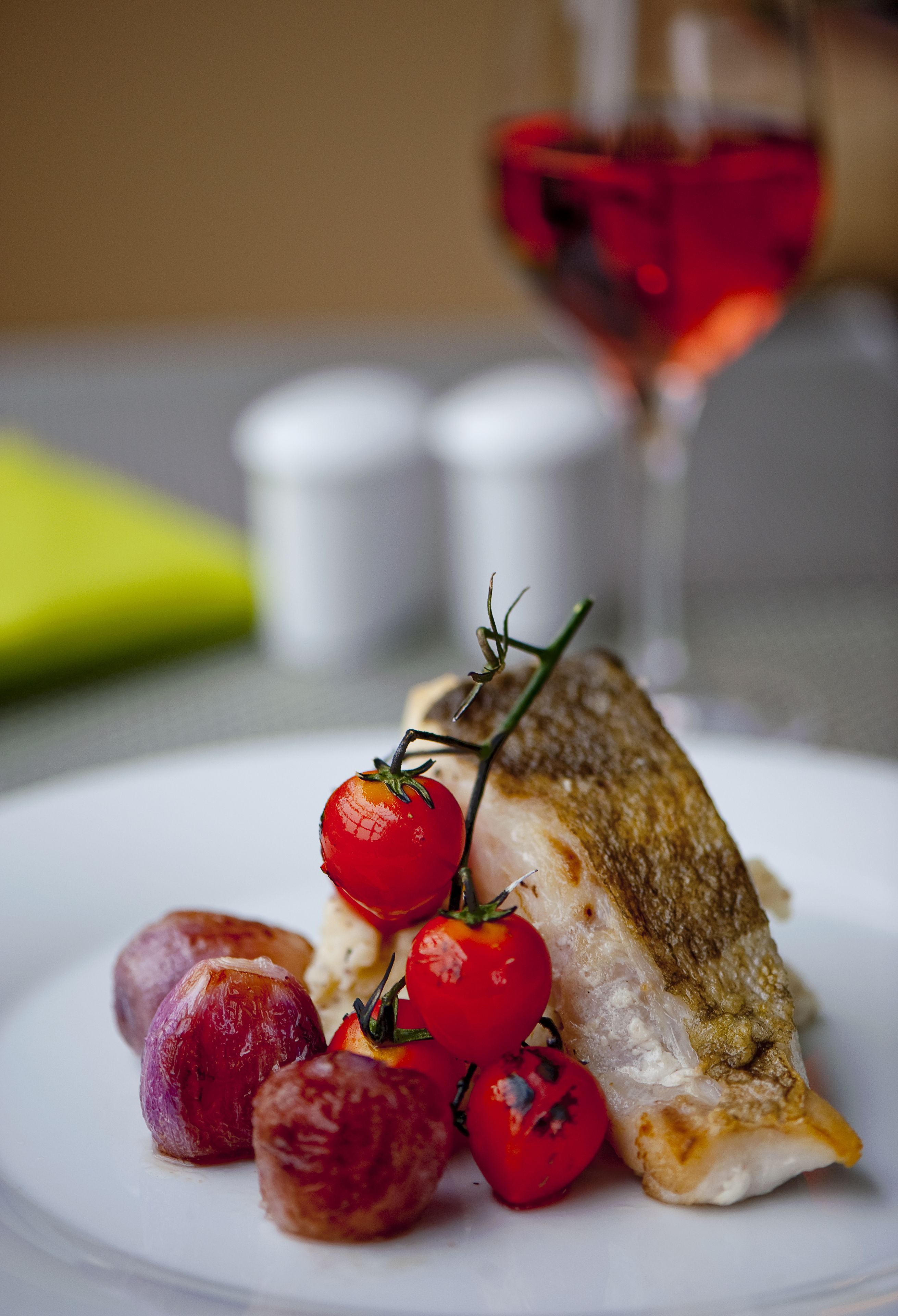 Un plat amb un peix, tomàquets cherries i petites rondalles, possiblement patates, damunt d'un plat blanc.