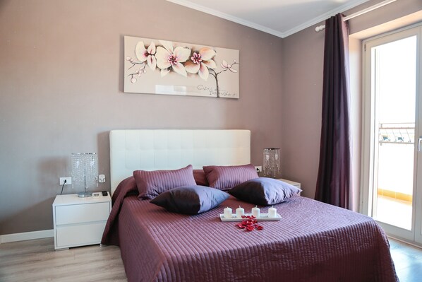 A bedroom with a large bed, a nightstand, a wall-mounted lamp, and a window with curtains.