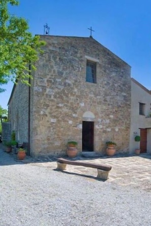 Un edificio in pietra con una croce sulla sommità, una piccola finestra e un portone. Davanti all'edificio c'è una panchina.