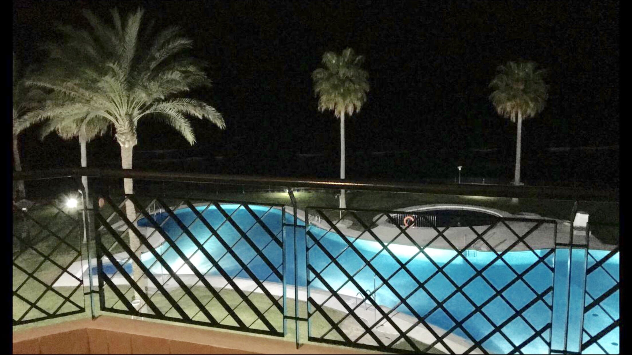 Vista notturna di una piscina con recinzione metallica, palme e una strada all'orizzonte.