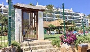 Una entrada protegida con puerta de madera, escalones que conducen a un edificio con tejado de paja, y vistas a una zona residencial con edificios blancos y balcones.