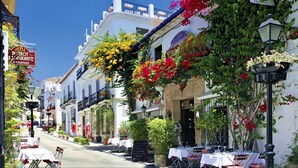 Una calle bordeada de edificios blancos, con mesas y sillas para exteriores y flores colgantes.