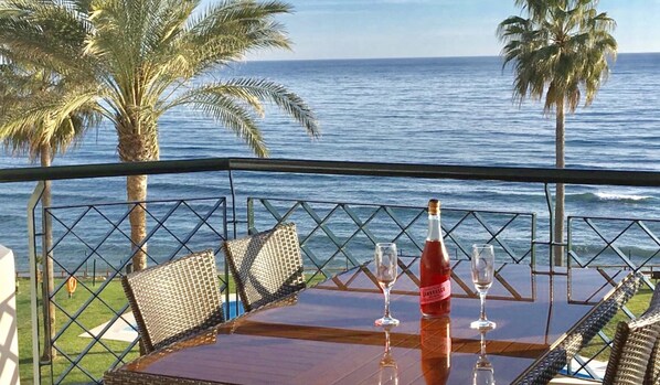 Mesa con una botella y dos vasos, con vista al océano y palmeras.