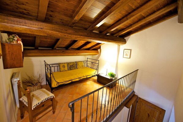 Un dormitorio tipo loft con techo de madera, una cama con colcha amarilla, una silla con cojín a cuadros, una mesita con una planta y un cuadro en la pared.