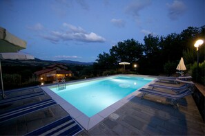 A rectangular swimming pool with a stone deck, surrounded by lounge chairs and umbrellas, with a view of hills and a small building in the background.