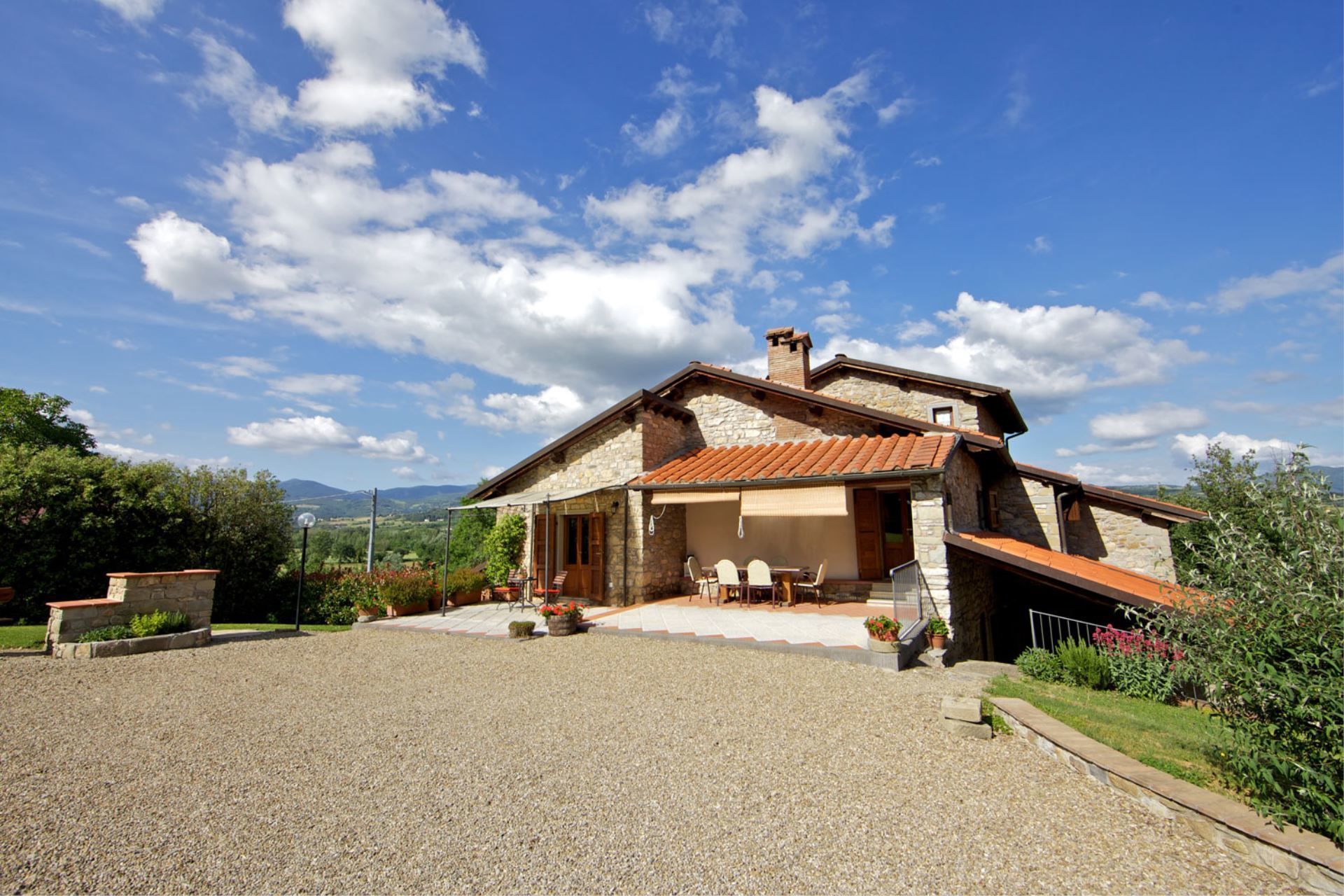 A stone house with a tiled roof, a covered patio with outdoor furniture, and a gravel driveway.