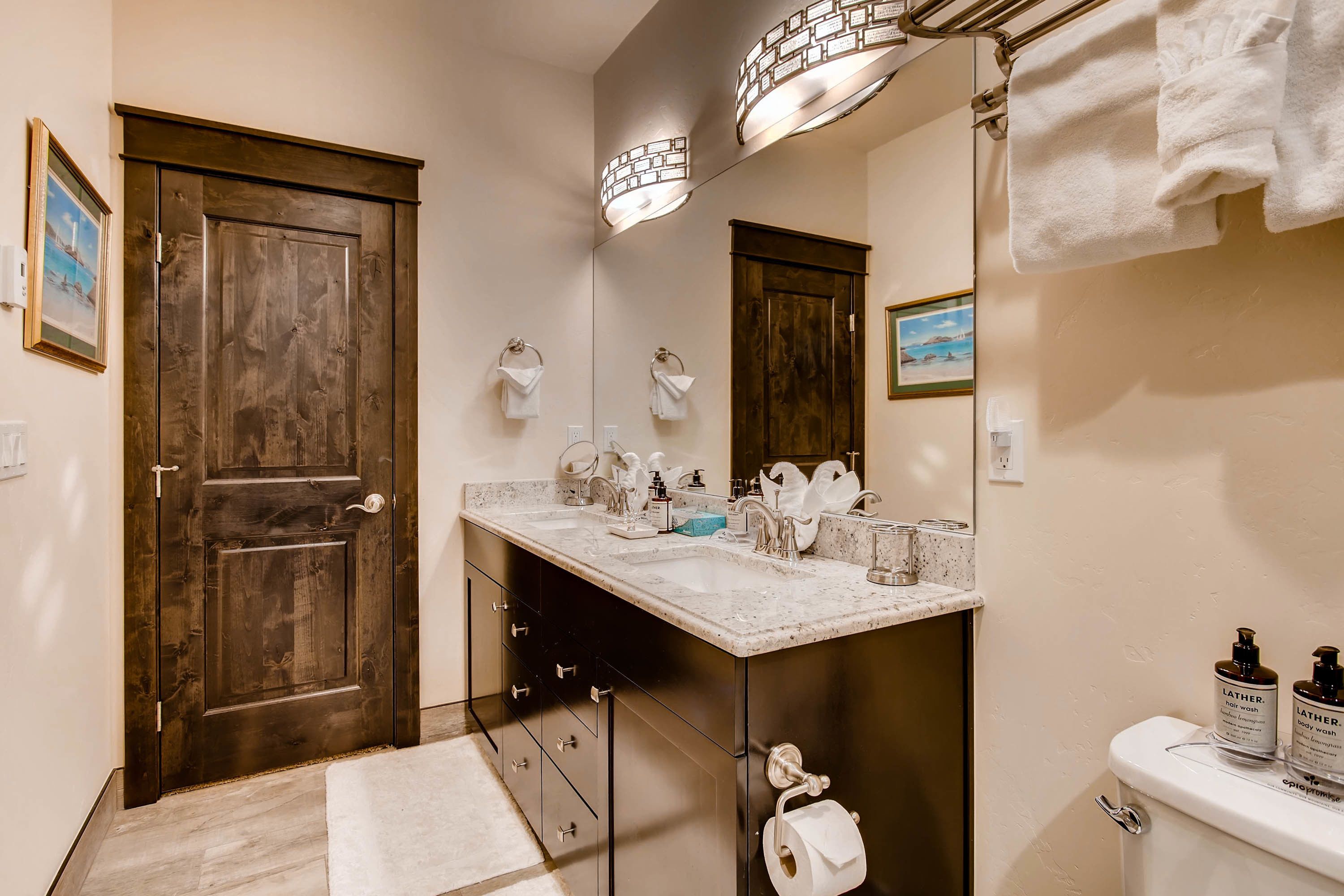 A bathroom with a wooden door, a sink with a mirror, and two bottles of Lather soap on the counter.