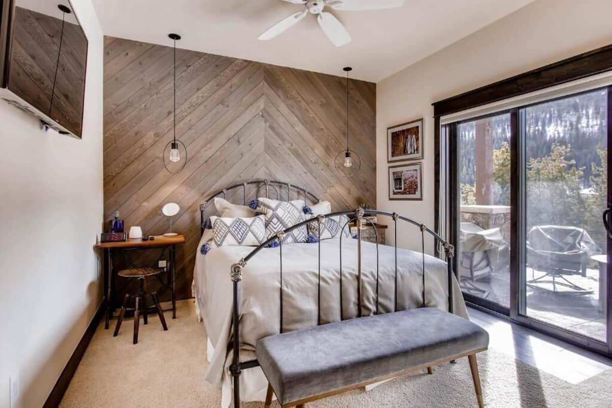 Une chambre avec un mur en bois à motifs chevrons, un lit avec un cadre en métal, un banc, un bureau avec une chaise et une grande fenêtre donnant sur des arbres.