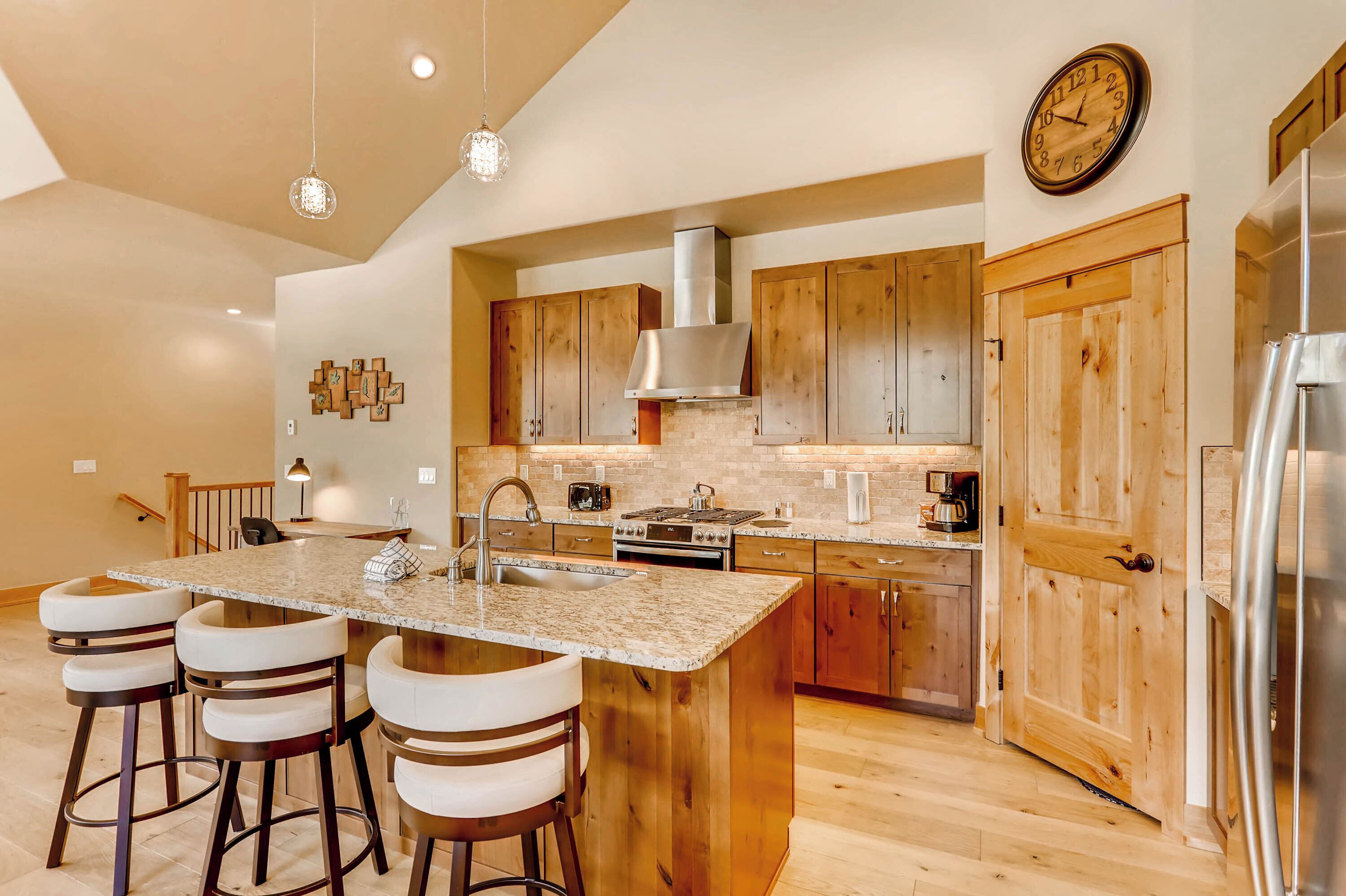 A modern kitchen with a central island, wooden cabinets, stainless steel appliances, and a clock on the wall.