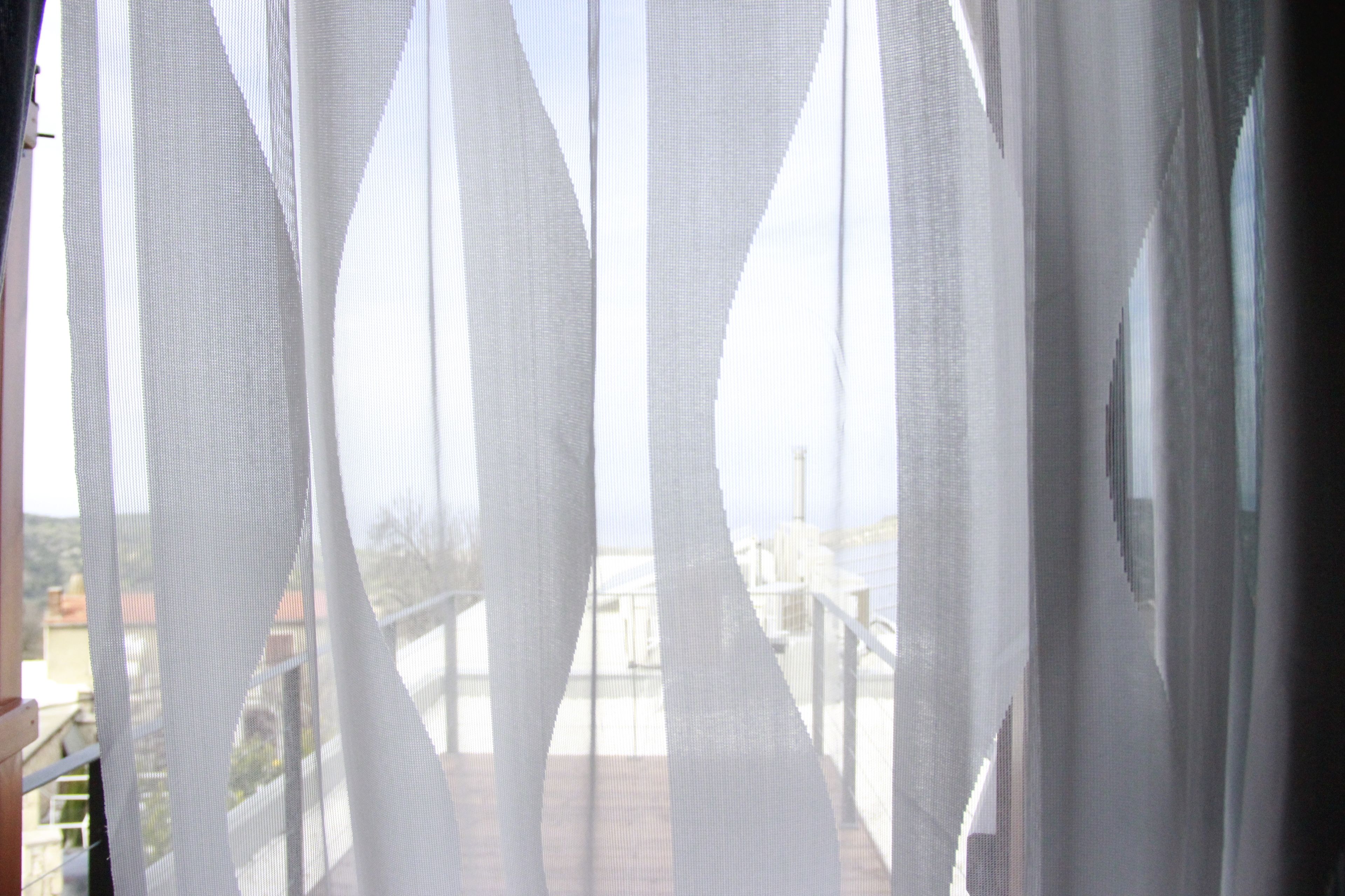 A balcony with sheer white curtains, a railing, and a view of buildings in the distance.