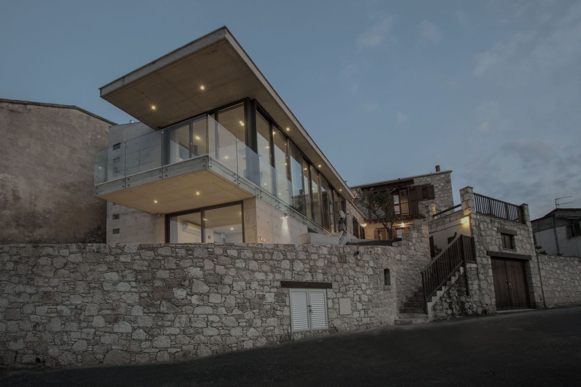 Une maison moderne à deux étages, dotée d’une façade en pierre, de grandes fenêtres et d’un balcon.