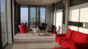 A modern living room with a red sofa, a red armchair, and a black armchair. There is a glass door leading to a balcony with a view of the city. - Marmaras (Lysos)