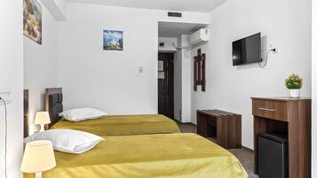 Hotellihuone, jossa on kaksi sänkyä, televisio, työpöytä, jääkaappi ja pieni huonekasvi.