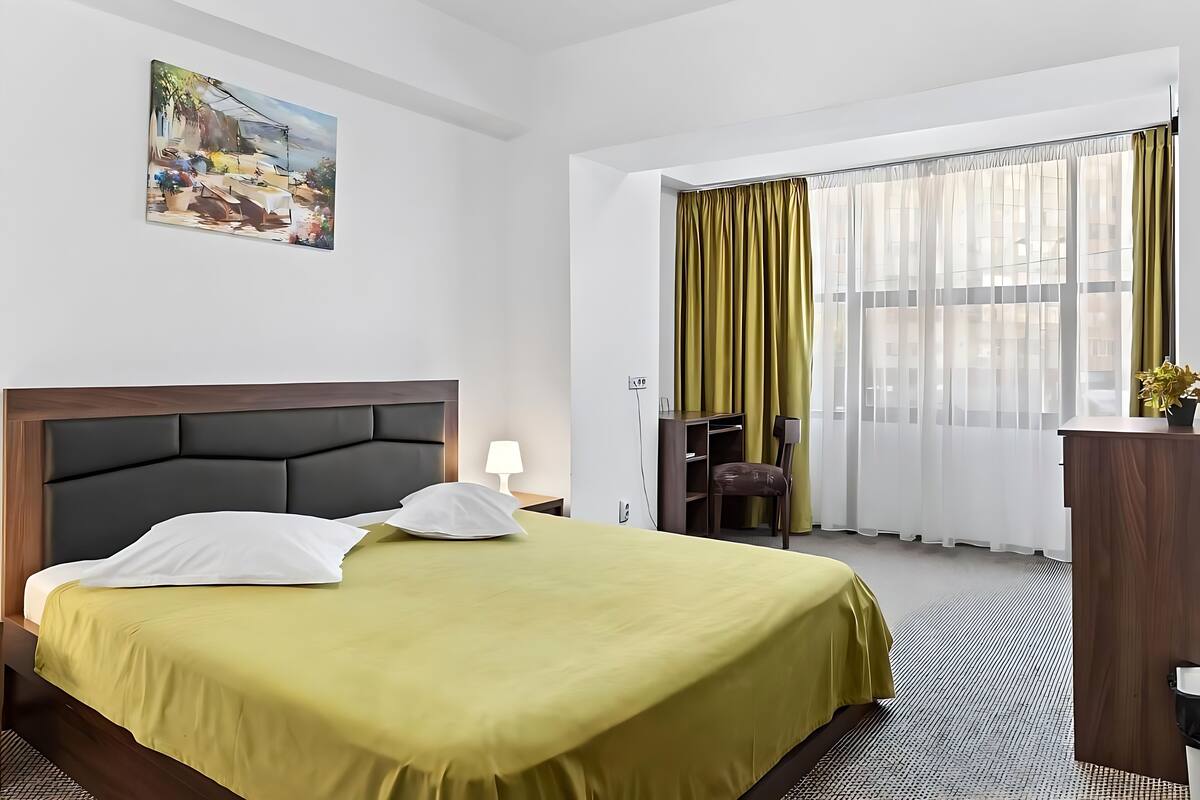 A hotel room with a bed, a desk, a chair, a lamp, and a window with curtains.
