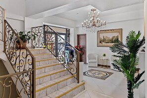 Una escalera con barandillas metálicas ornamentadas, un candelabro y un cuadro grande en la pared.