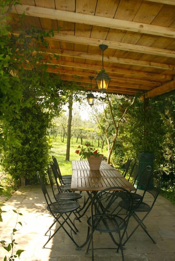 Eine überdachte Terrasse mit einem Holztisch und Metallstühlen, umgeben von viel Grün und Blick auf einen Garten.