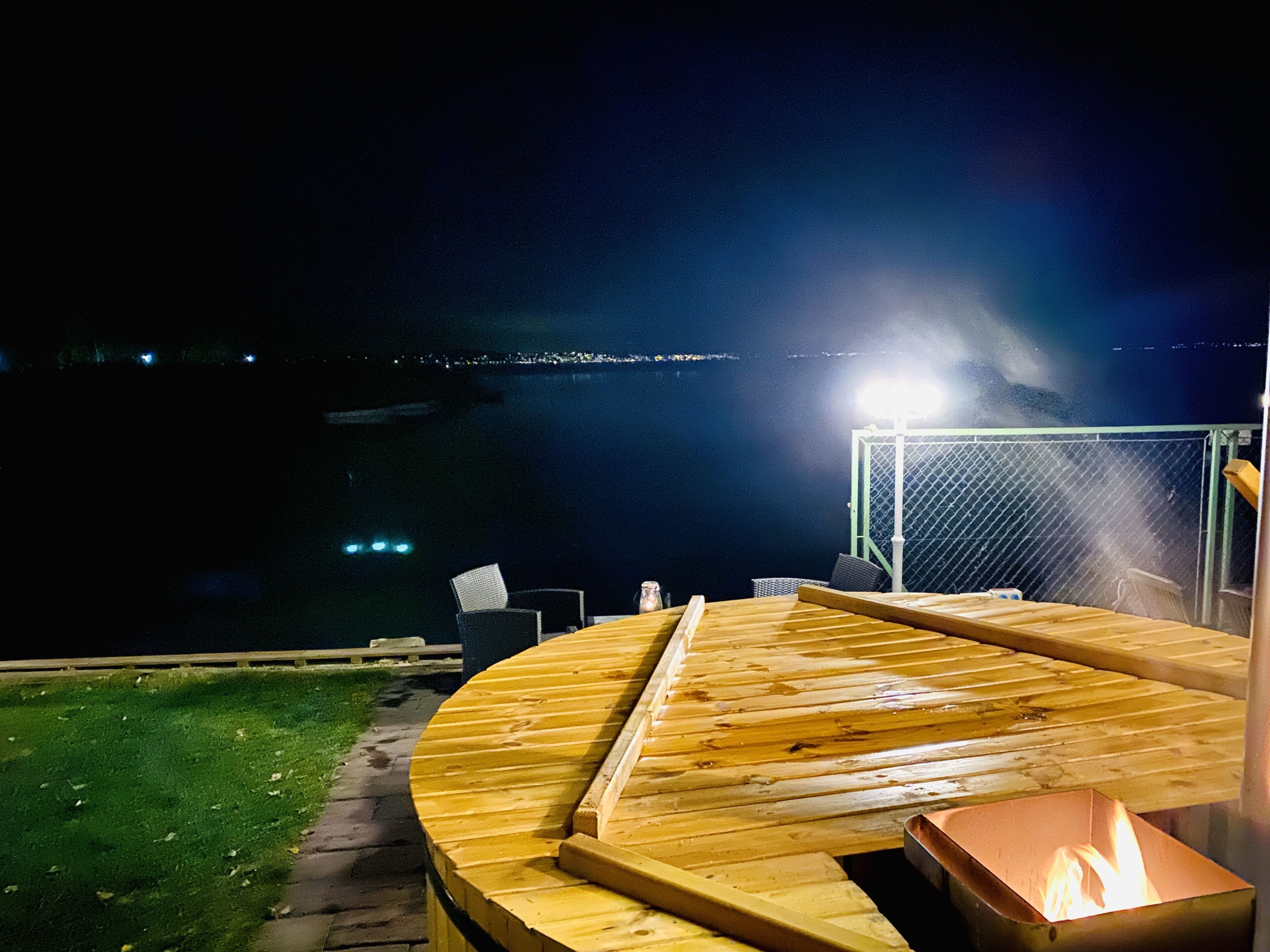 Una terraza de madera con un hoyo de fuego y zona de estar con vistas a un cuerpo de agua al caer la noche.