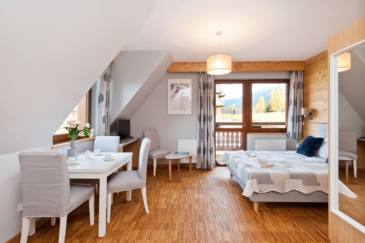 Une pièce moderne et bien éclairée, dotée d’un plancher en bois, d’un lit, d’une table à manger avec des chaises, d’une petite table ornée d’un vase de fleurs et offrant une vue sur les montagnes par la fenêtre.