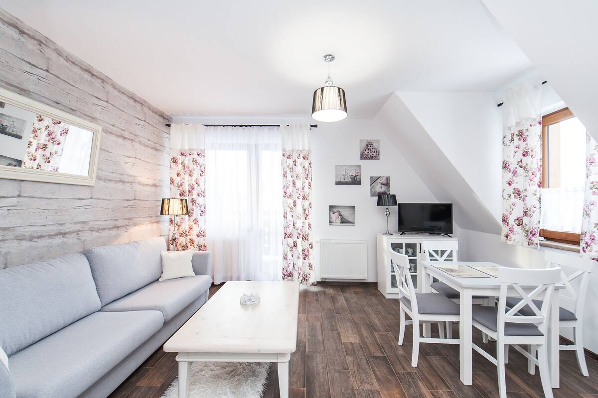 Un salon moderne comprenant un canapé, une table basse, une table à manger et des chaises. On y trouve également un téléviseur, un meuble blanc et une fenêtre ornée de rideaux à motifs floraux.