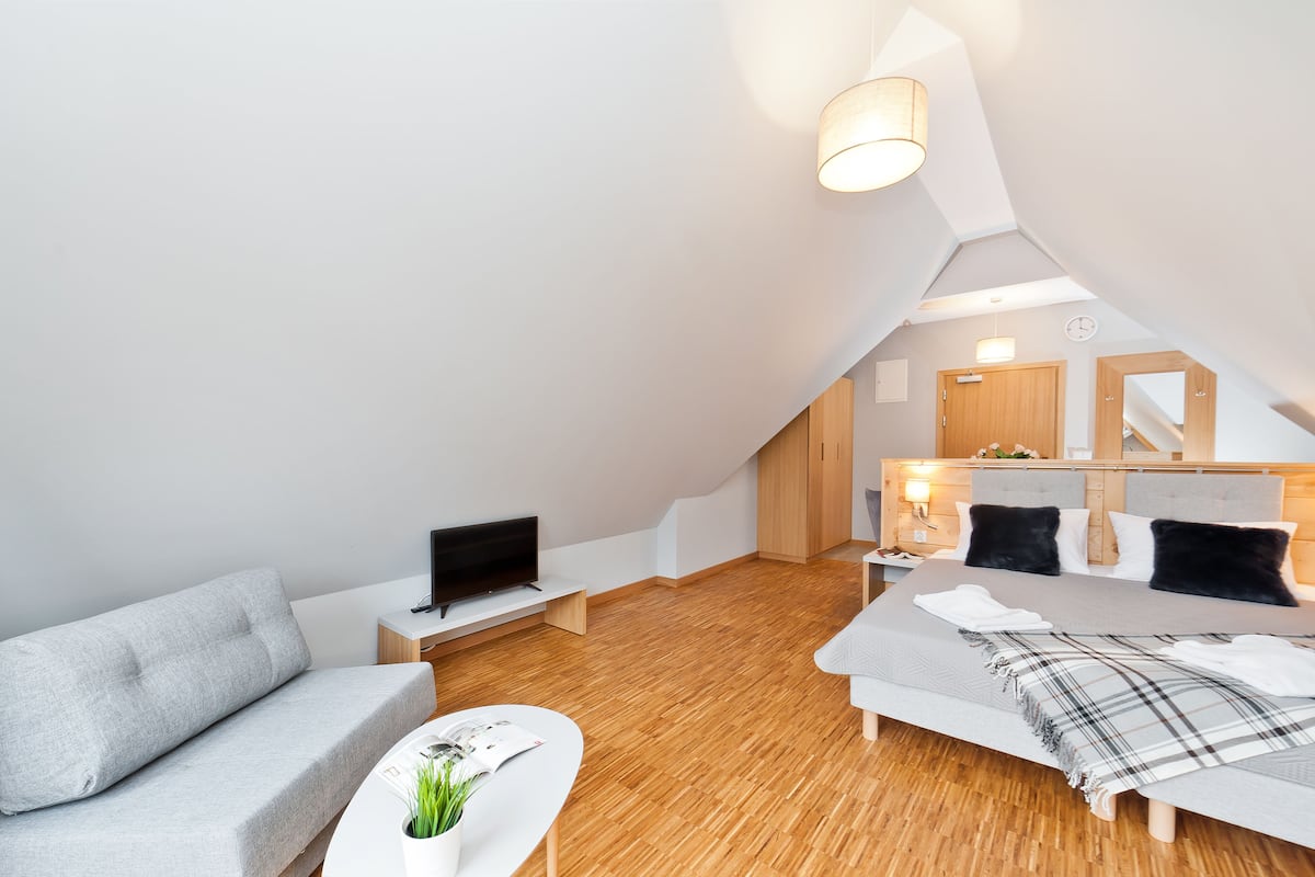 Une chambre d’hôtel moderne avec un plafond de style industriel, un lit recouvert d’une couverture grise, un canapé gris, un téléviseur à écran plat et une petite table sur laquelle est posée une plante.