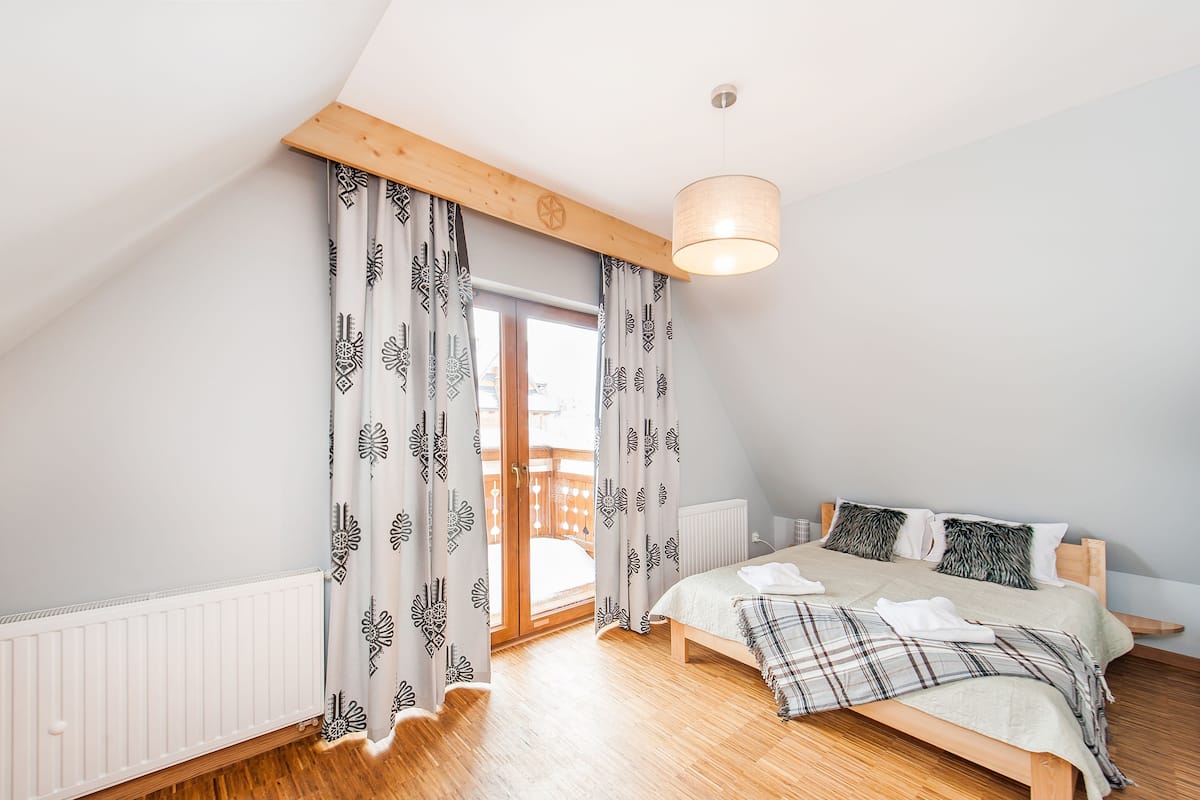 Une chambre avec un plafond en bois, un lit recouvert d’une couverture à carreaux, un radiateur blanc et des rideaux à motifs.