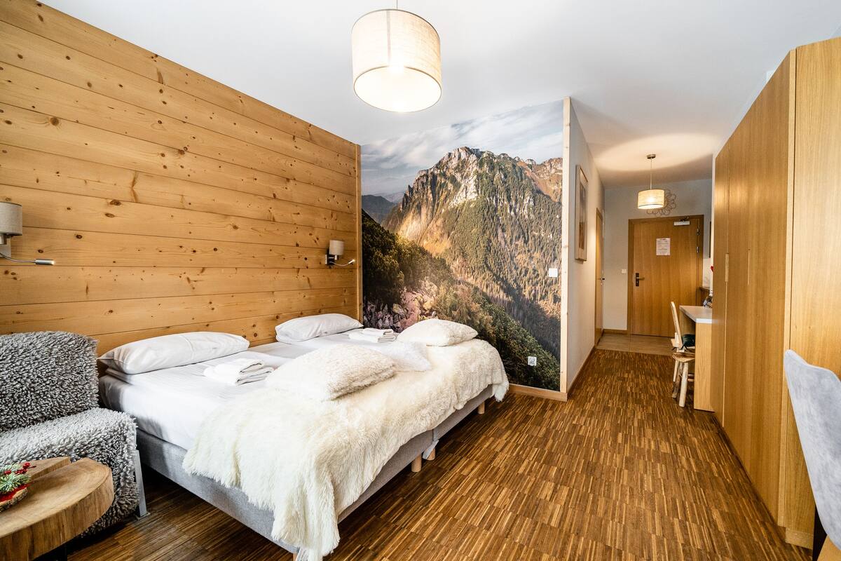 Une chambre d’hôtel avec des murs en bois, un lit avec une literie blanche, une vue sur les montagnes, un plancher en bois et un petit bureau avec une chaise.