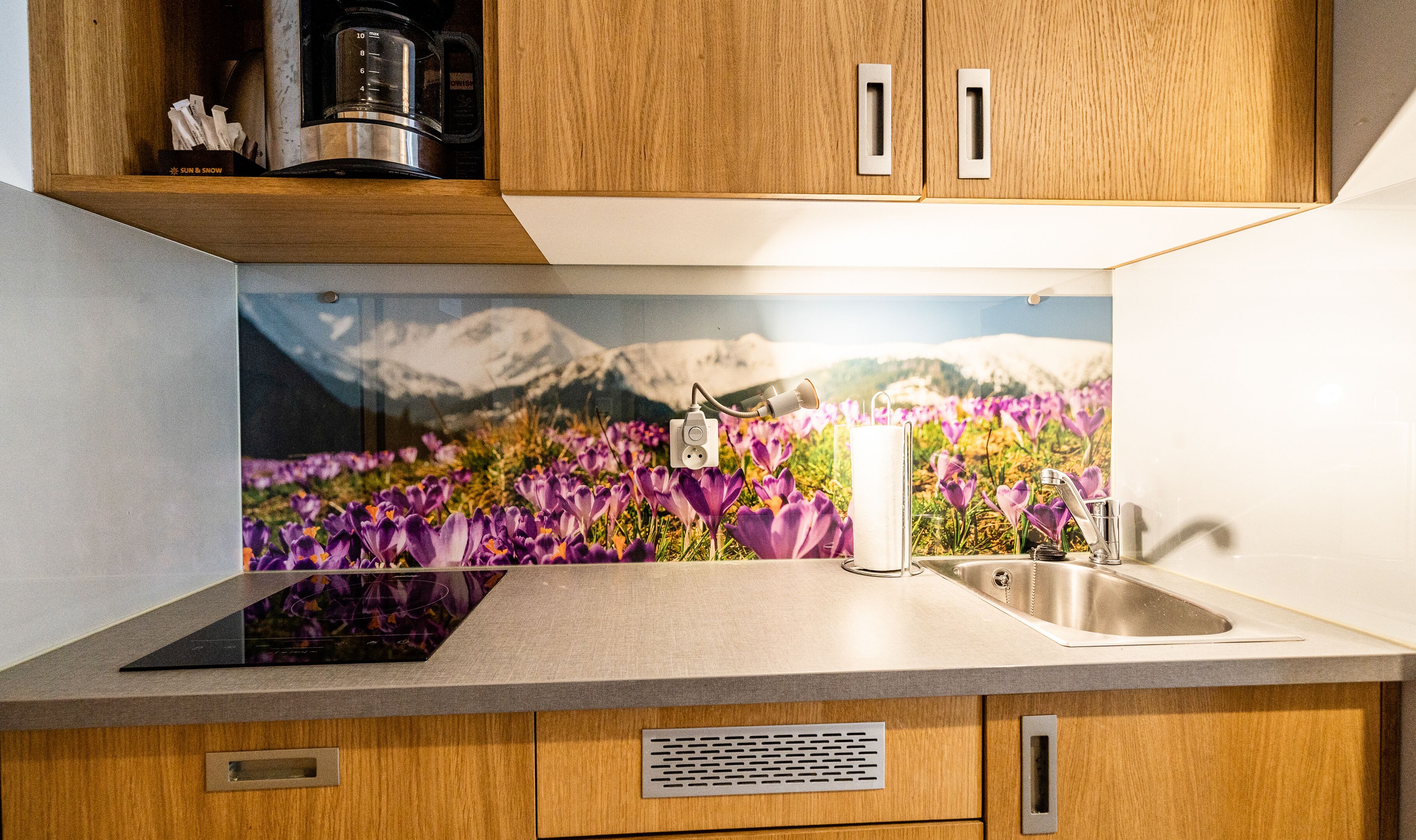 A kitchen with wooden cabinets, a stainless steel sink, and a backsplash with a mountain and flower scene.