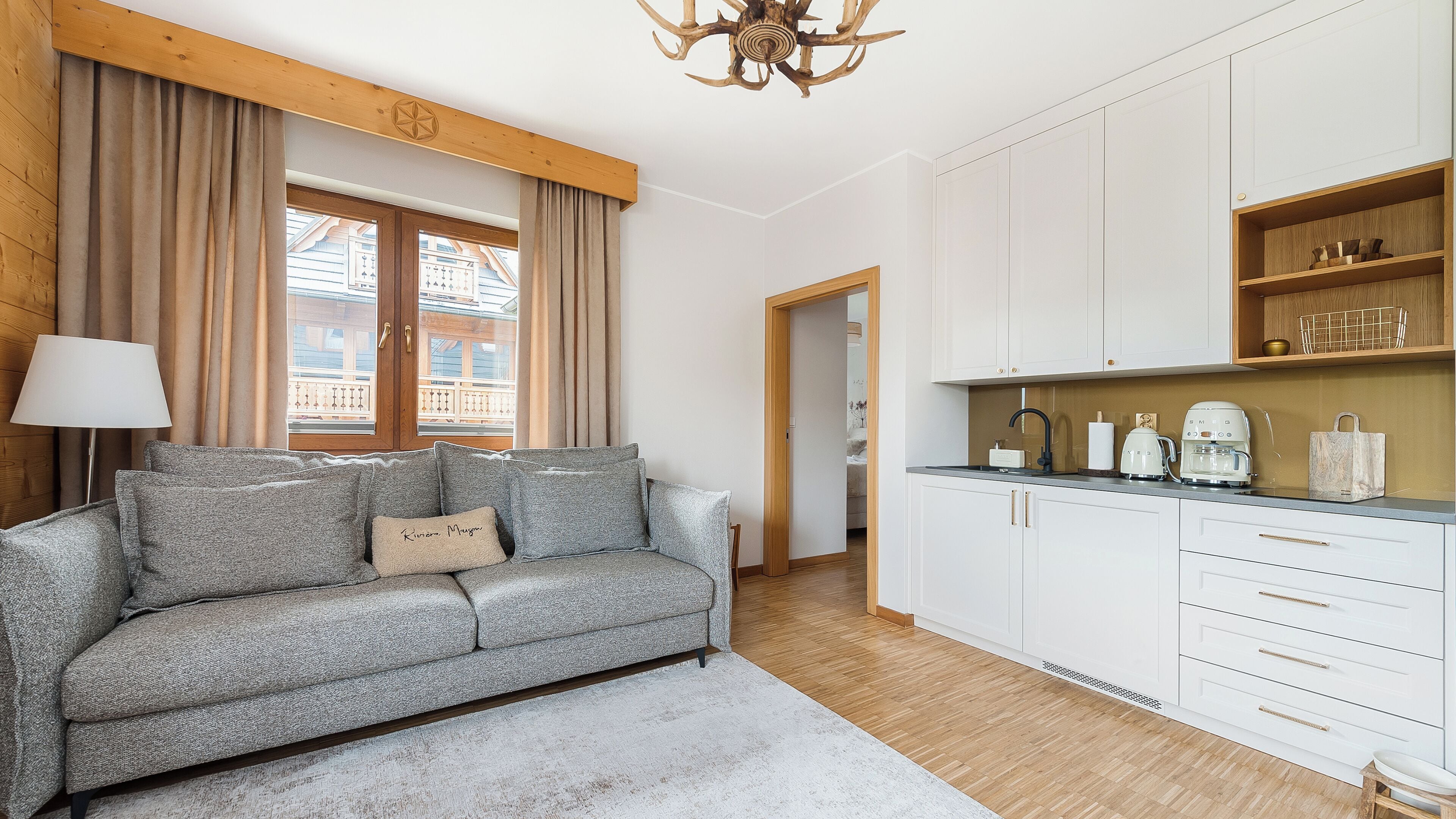 A modern living room with a grey sofa, a wooden ceiling, and a kitchen area with white cabinets and a coffee machine.