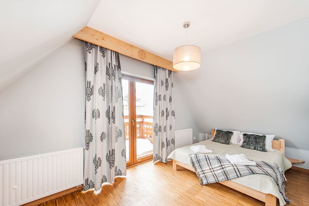 Une chambre avec un plafond en pente, des poutres en bois, un lit recouvert d’une couverture à carreaux et des rideaux à motifs.