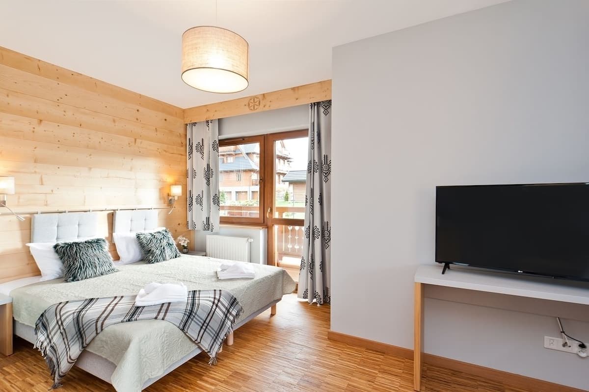 Une chambre à coucher avec un lit, un téléviseur posé sur un meuble, des murs en bois, une fenêtre avec des rideaux et un luminaire au plafond.
