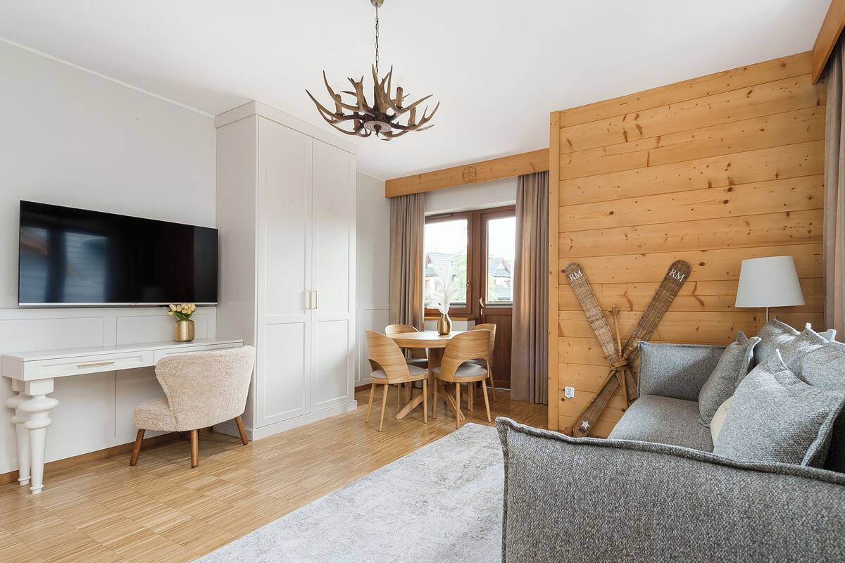 Un salon moderne avec un mur en bois, un téléviseur fixé au mur, une table à manger avec des chaises et un canapé agrémenté de coussins.