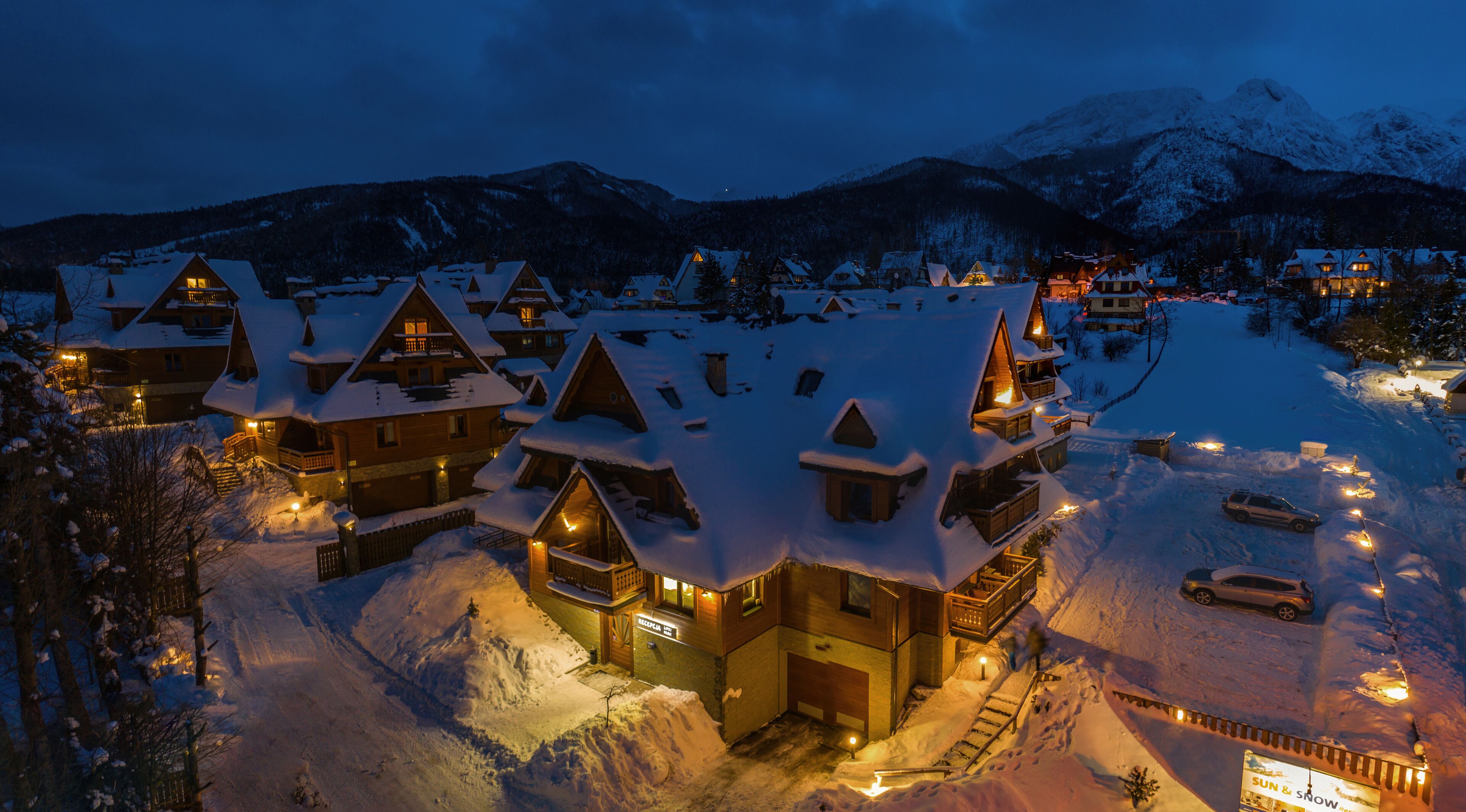 Un albergue cubierto de nieve con varias edificaciones, un telesilla y un telón de fondo montañoso.
