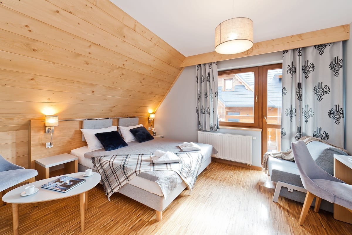 Une chambre confortable avec un plafond en bois, un lit recouvert d’une couverture à carreaux, une petite table avec un magazine et une chaise.