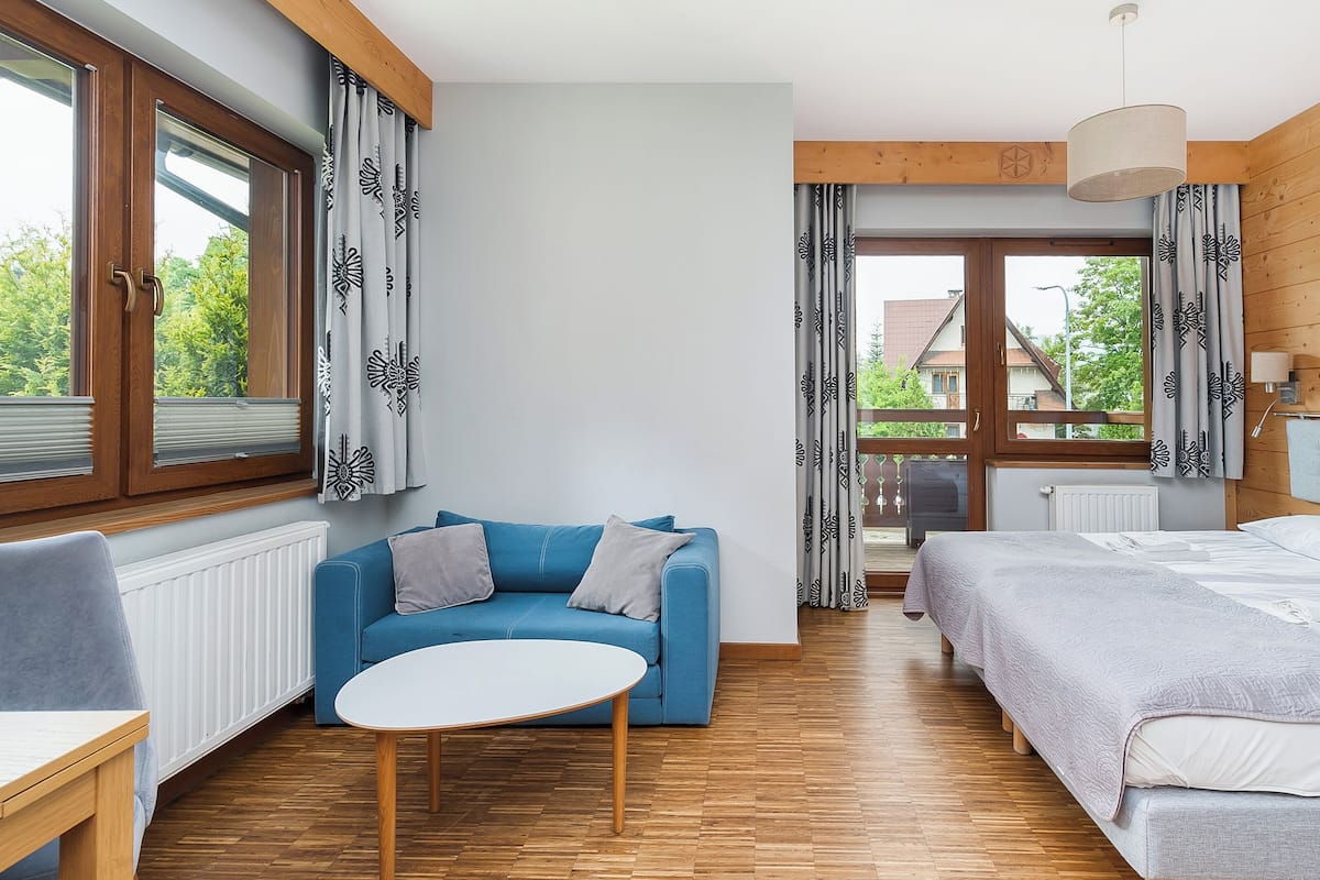 Une pièce avec un canapé bleu, un lit, une petite table ronde, un plancher en bois et de grandes fenêtres ornées de rideaux à motifs.