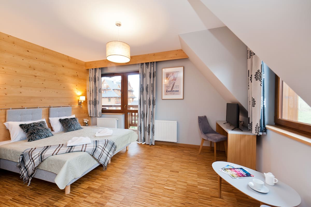 Une chambre moderne avec un plancher en bois, un lit recouvert d’une couette grise et blanche, une table de chevet avec une lampe et une fenêtre avec des rideaux.