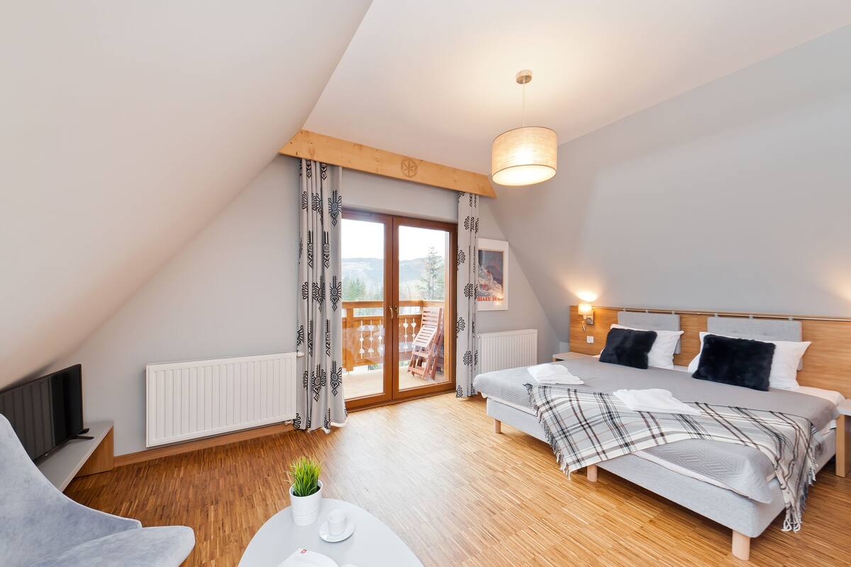 Une chambre moderne avec un lit, une télévision, une petite table avec une plante et un balcon doté d’une terrasse en bois.