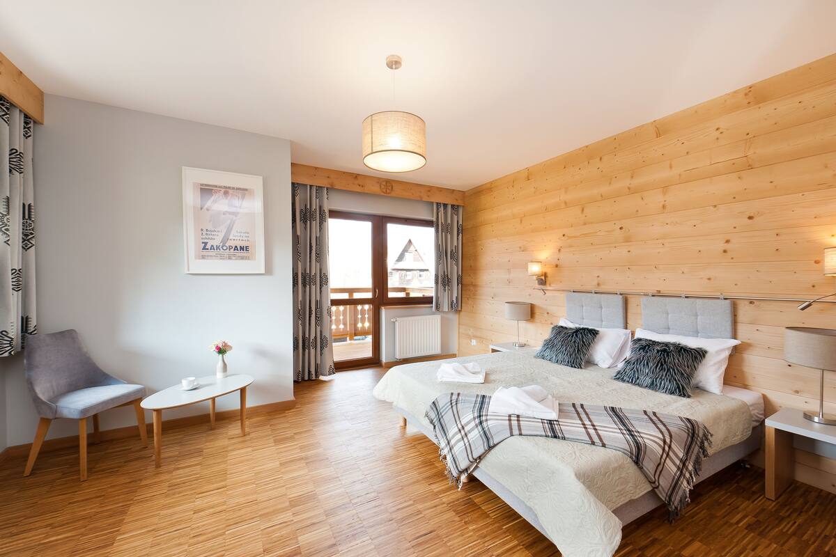 Une chambre avec un plancher en bois, un lit doté d’une tête de lit grise, une table de chevet avec une lampe et une fenêtre avec des rideaux.