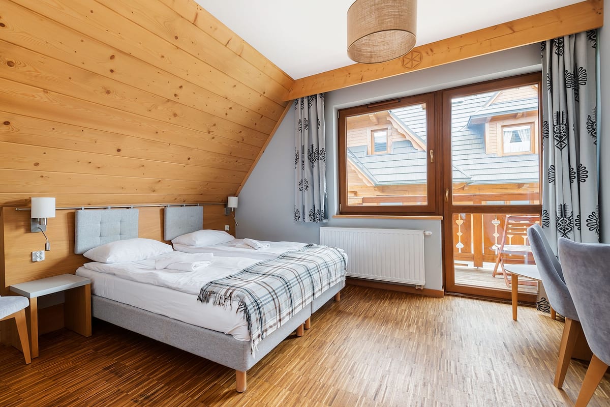 Une chambre avec un plafond en bois, un lit recouvert d’une couverture à carreaux, une chaise, une table de chevet et une fenêtre avec des rideaux.