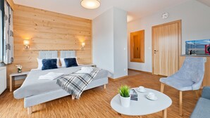 Une chambre d’hôtel moderne avec un grand lit, une table basse ronde, une chaise et une petite table sur laquelle se trouvent une plante et des tasses.