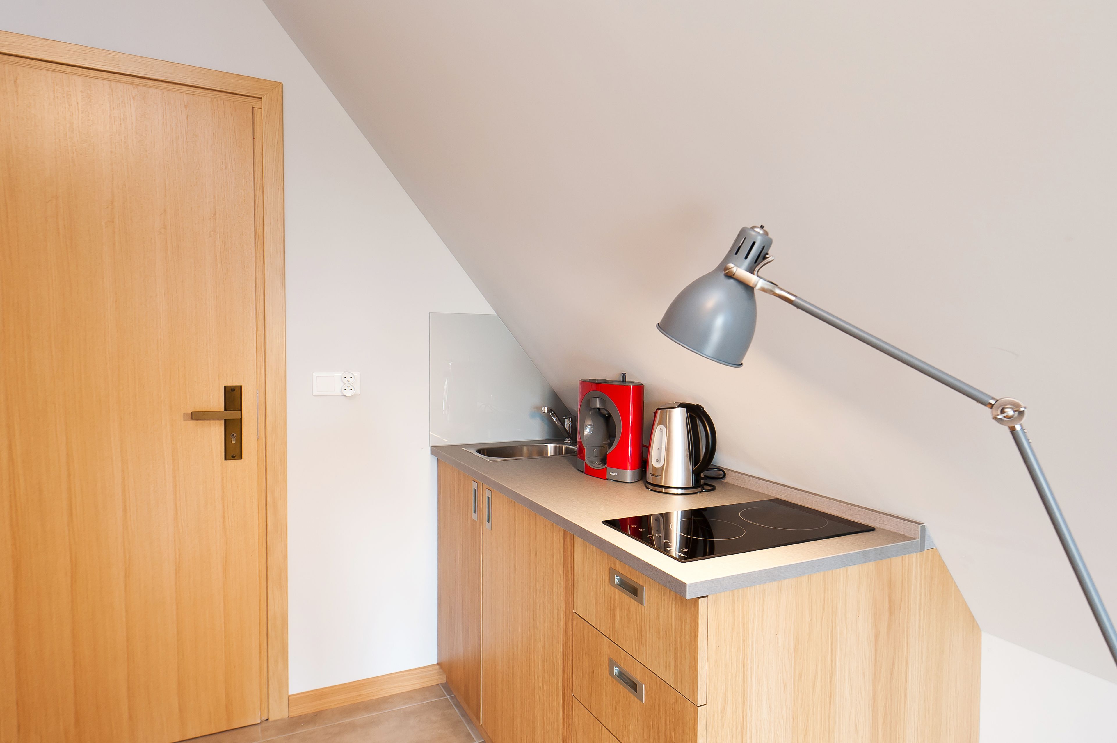 Una kitchenette pequeña con un mueble de madera, un lavamanos, una tetera roja y una lámpara moderna.