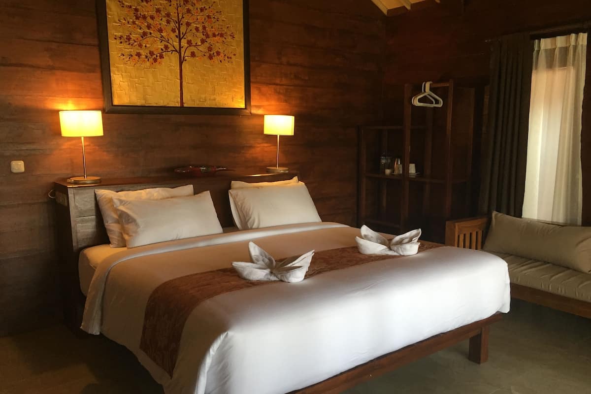 A neatly made bed with white linens and decorative pillows, two bedside lamps, a wooden headboard, a wooden wardrobe, and a window with curtains.