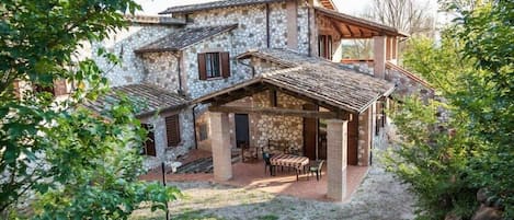 A stone house with a patio area, a covered outdoor seating space, and a view of the surrounding greenery.