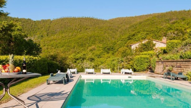 Una piscina rectangular con agua azul cristalina, rodeada por un solárium empedrado. Varias sillas de descanso blancas y una mesa con una botella de vino y fruta. El entorno es al aire libre, con una ladera verde y exuberante al fondo.