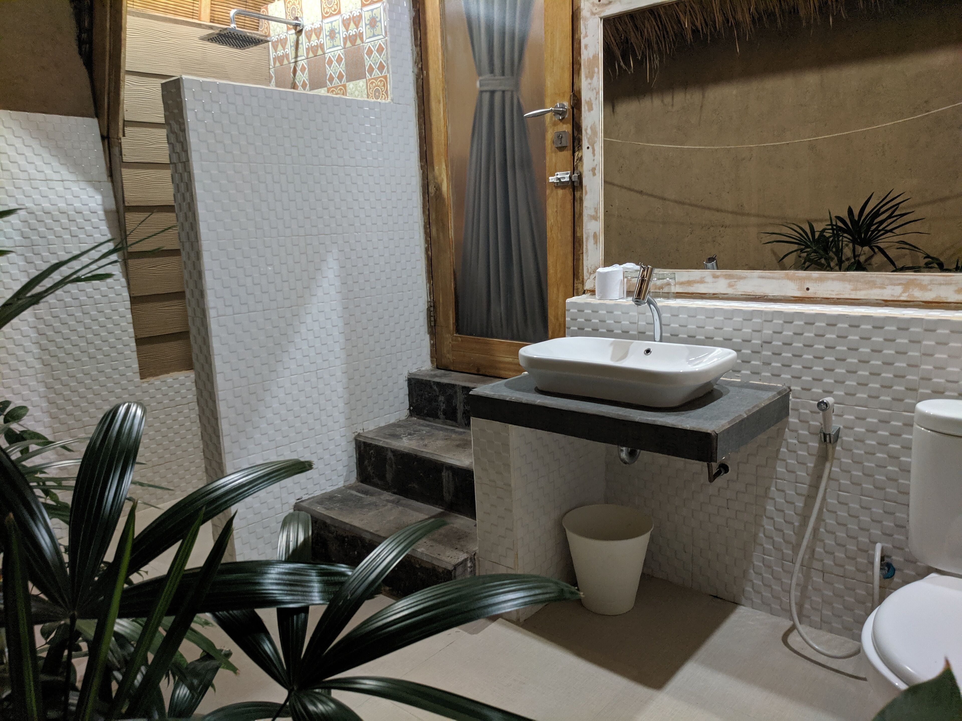 Een badkamer met een witte wastafel, een bad en een toilet. Er zijn treden die naar het bad leiden, en een plant is in de voorgrond te zien.