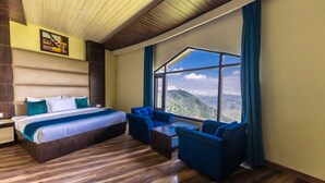 Habitación de hotel con una cama grande, dos sillones azules, una mesita y un ventanal con vistas a la montaña.