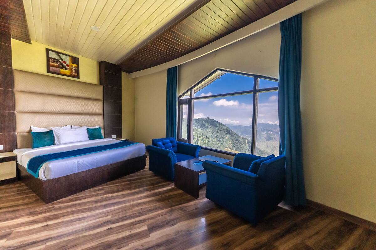 A hotel room with a large bed, two blue armchairs, a small table, and a large window offering a mountain view.