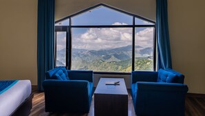 Habitación con un amplio ventanal que ofrece vistas a la montaña, dos sillones azules, una mesita y una cama.