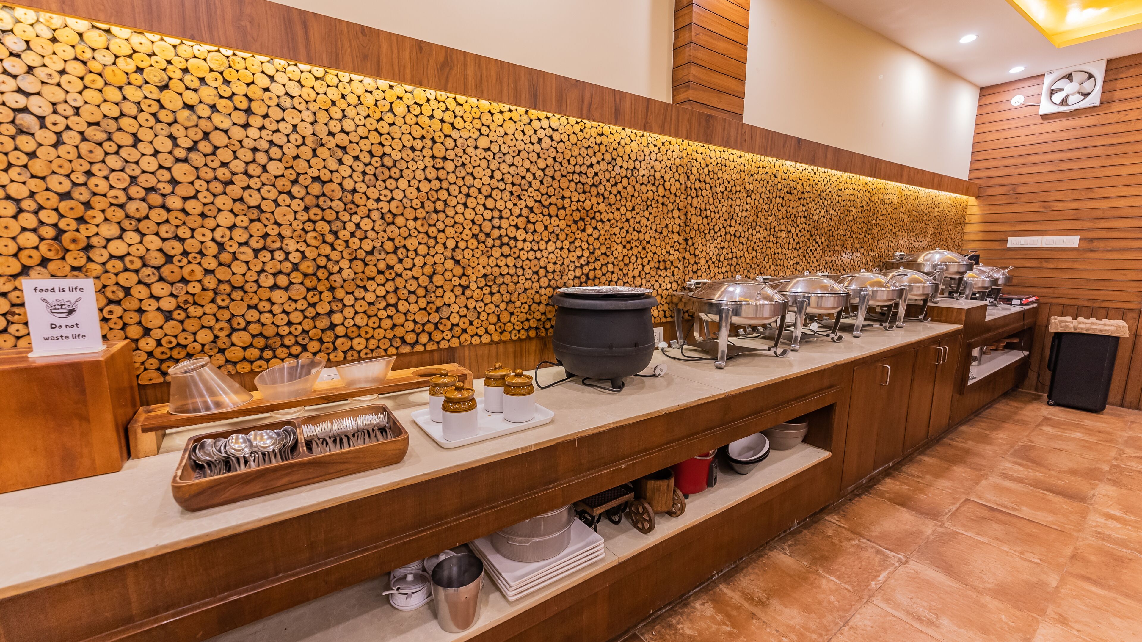 A buffet setup with wooden wall decor, buffet trolleys, and a sign with a food-related message.