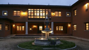 A building with a central fountain, multiple windows with lights, and a well-maintained lawn.