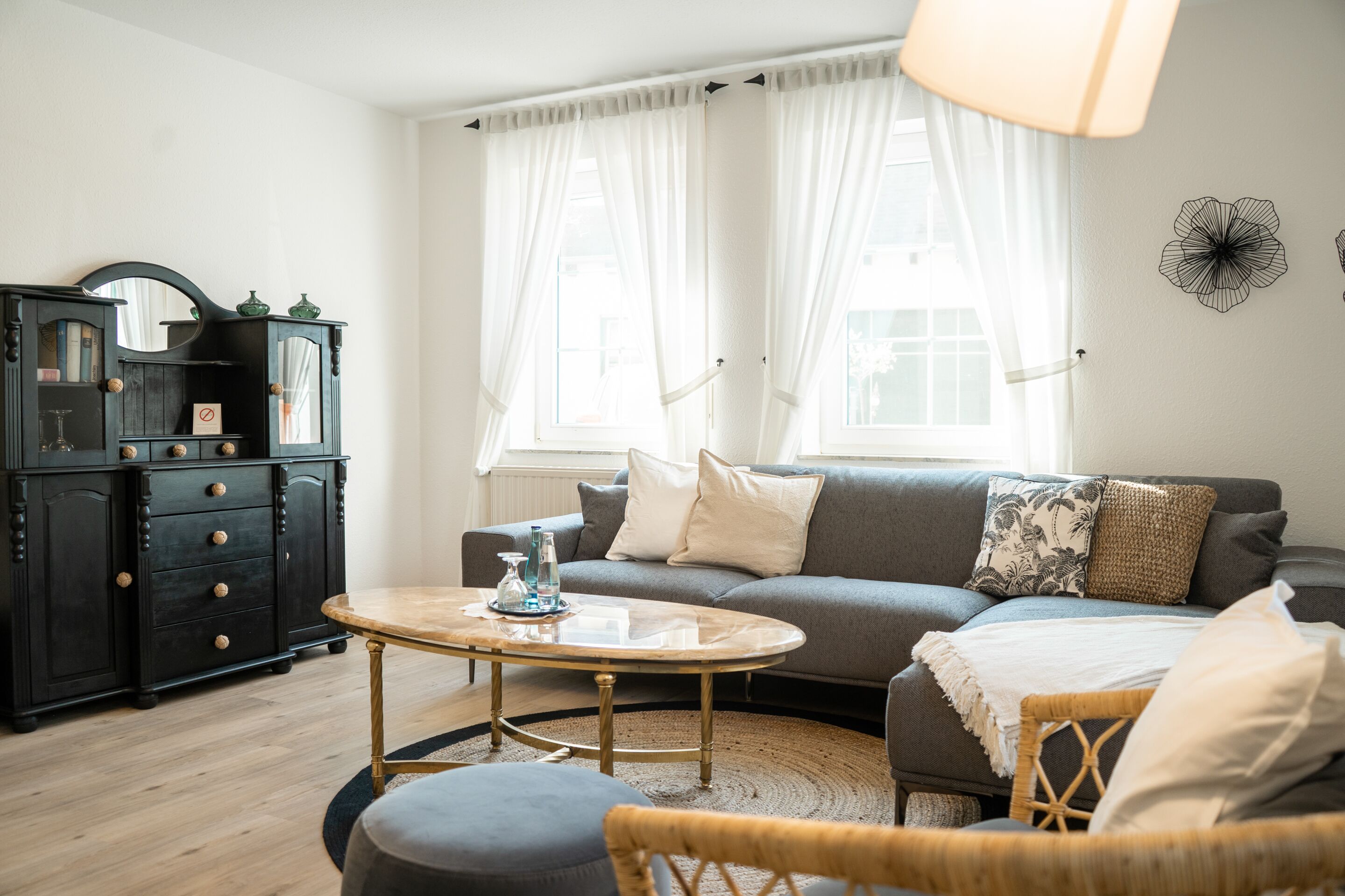 A living room with a sofa, a coffee table, a wicker chair, a black cabinet with drawers, and a large window with sheer curtains.